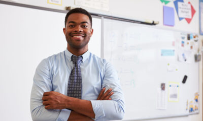 Male teacher stands in front of a white board in a classroom. He is smiling with his arms crossed.