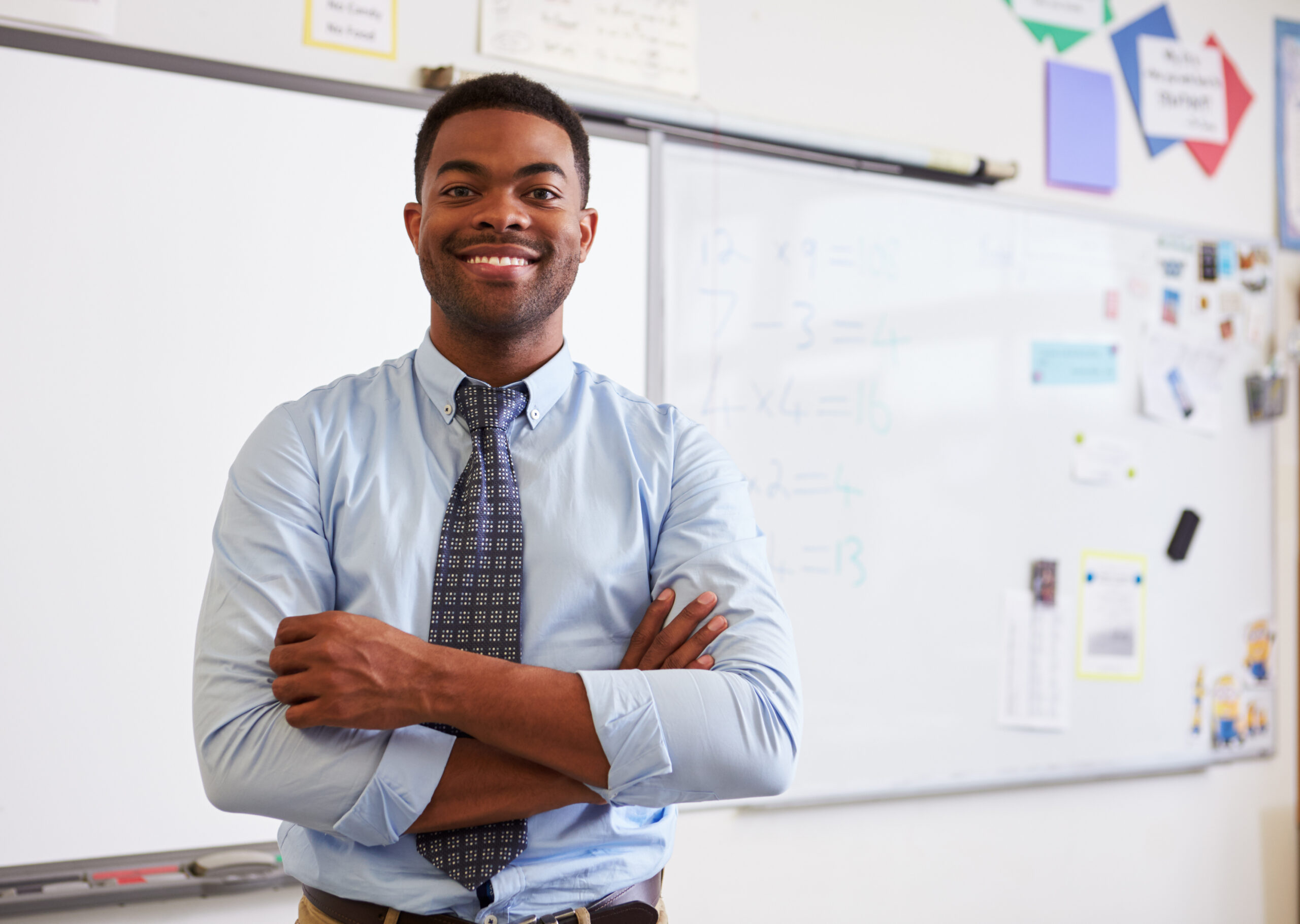 Male teacher stands in front of a white board in a classroom. He is smiling with his arms crossed.